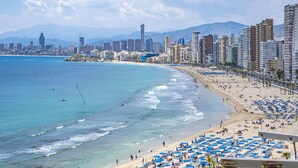 Un paisaje costero con una playa bordeada de sombrillas azules y diversos edificios, incluyendo apartamentos de gran altura y hoteles.