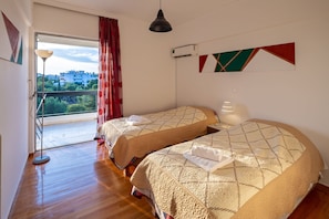 A hotel room with two beds, a balcony with red curtains, an air conditioning unit, and a view of buildings and trees.
