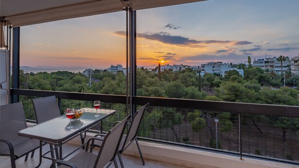 A balcony with a table and chairs, overlooking a cityscape during sunset. - Voula- Archer Amazing Sea View Apartment (Vari-Voula-Vouliagmeni)