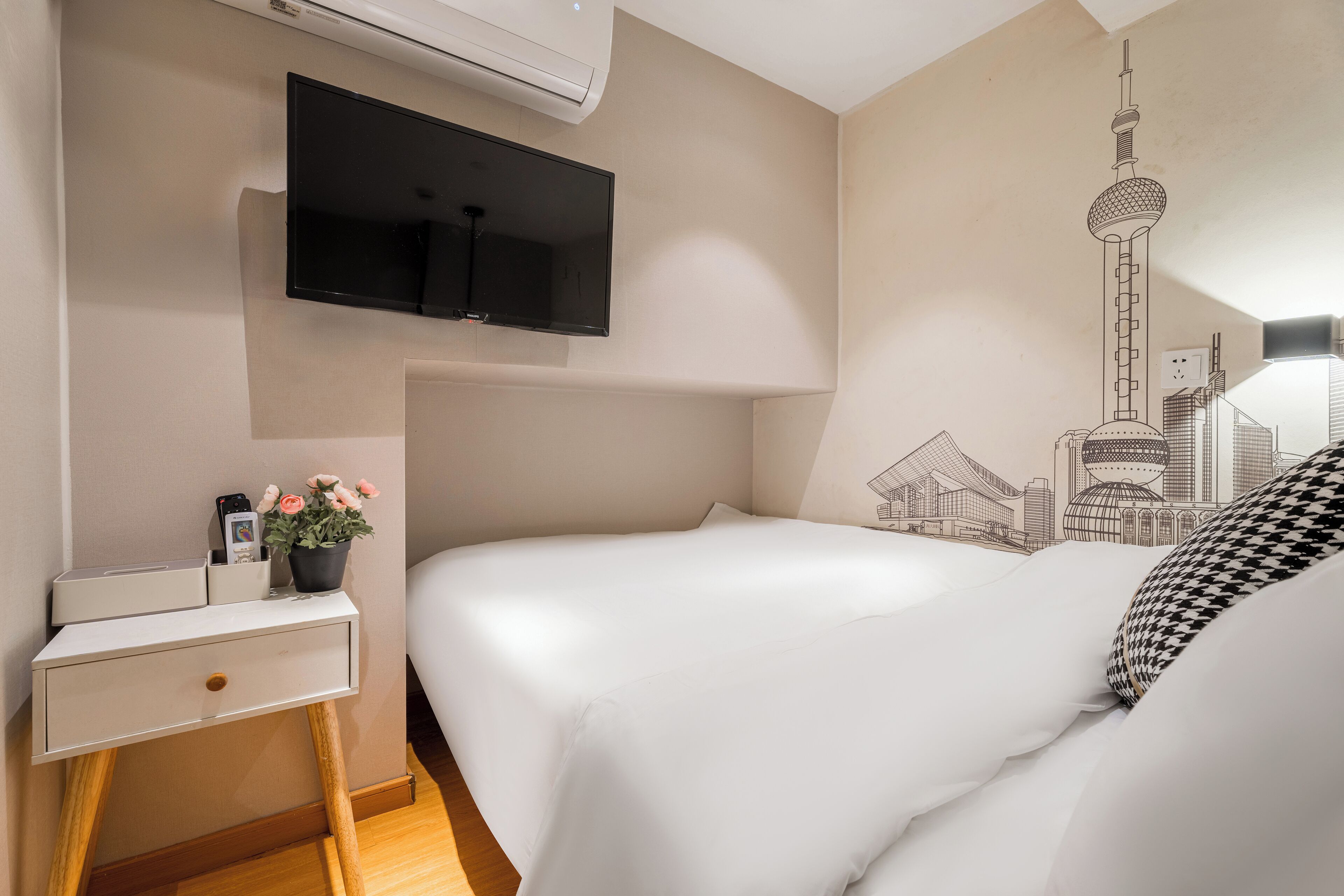 A hotel room with a bed, a bedside table, a TV mounted on the wall, and a wall mural of a cityscape.