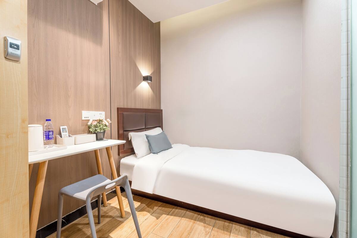 A hotel room with a bed, a desk with a chair, a small table, a water bottle, a phone, and a wall-mounted light fixture.