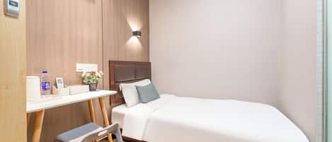 A hotel room with a bed, a desk with a chair, a small table, a water bottle, a phone, and a wall-mounted light fixture.