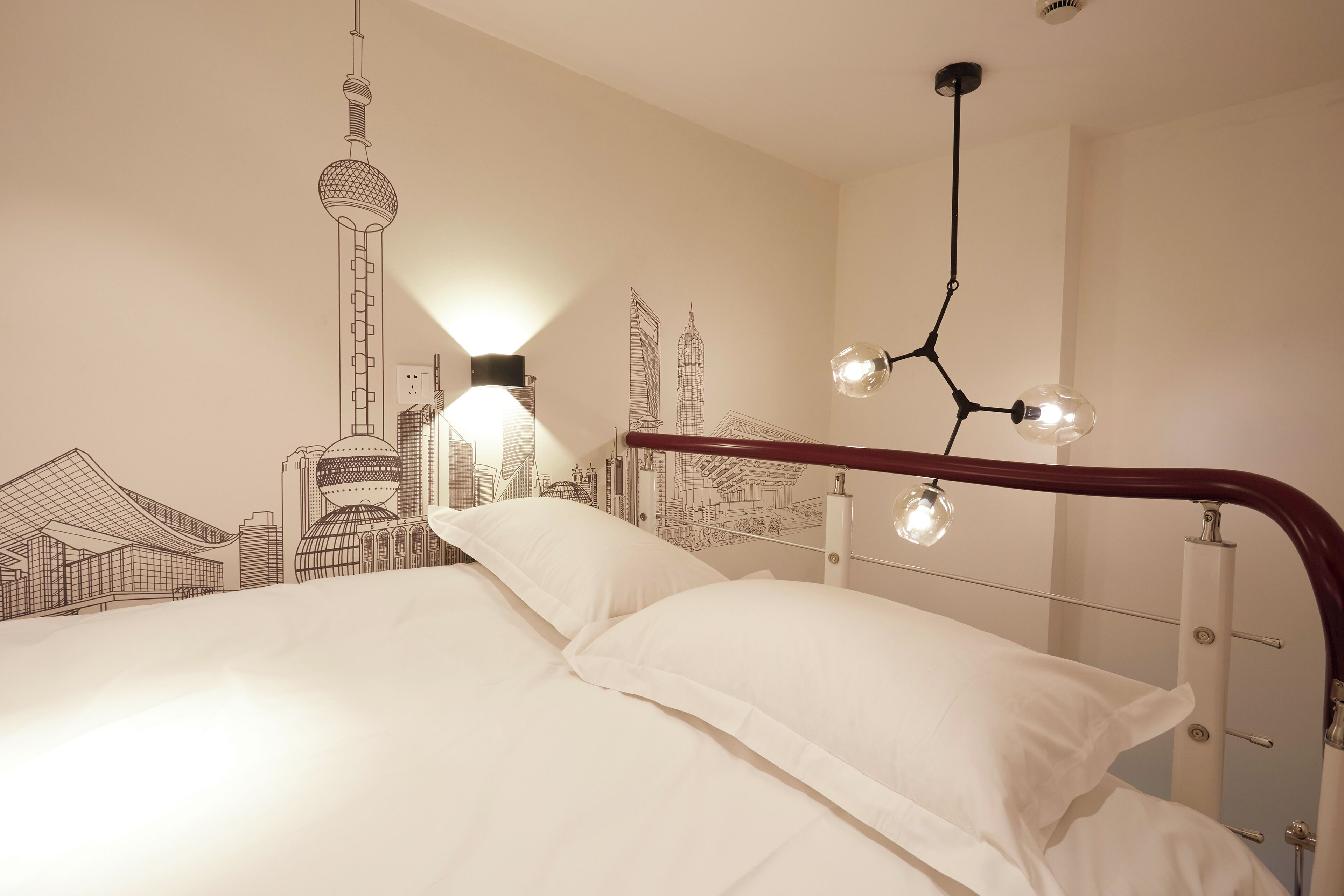 Een bed met witte lakens en een hoofdbord met een golvend ontwerp. Aan de muur een muurschildering van stadsgezichten en een hanglamp erboven.