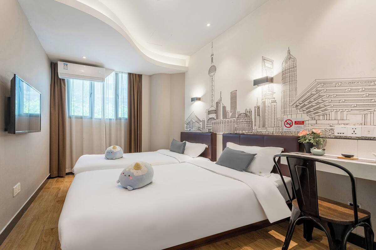A hotel room with two beds, a flat-screen TV, an air conditioning unit, a cityscape mural, and a desk with a chair.