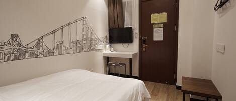 A hotel room with a bed, a desk with a chair, a TV mounted on the wall, and a door with a sign.