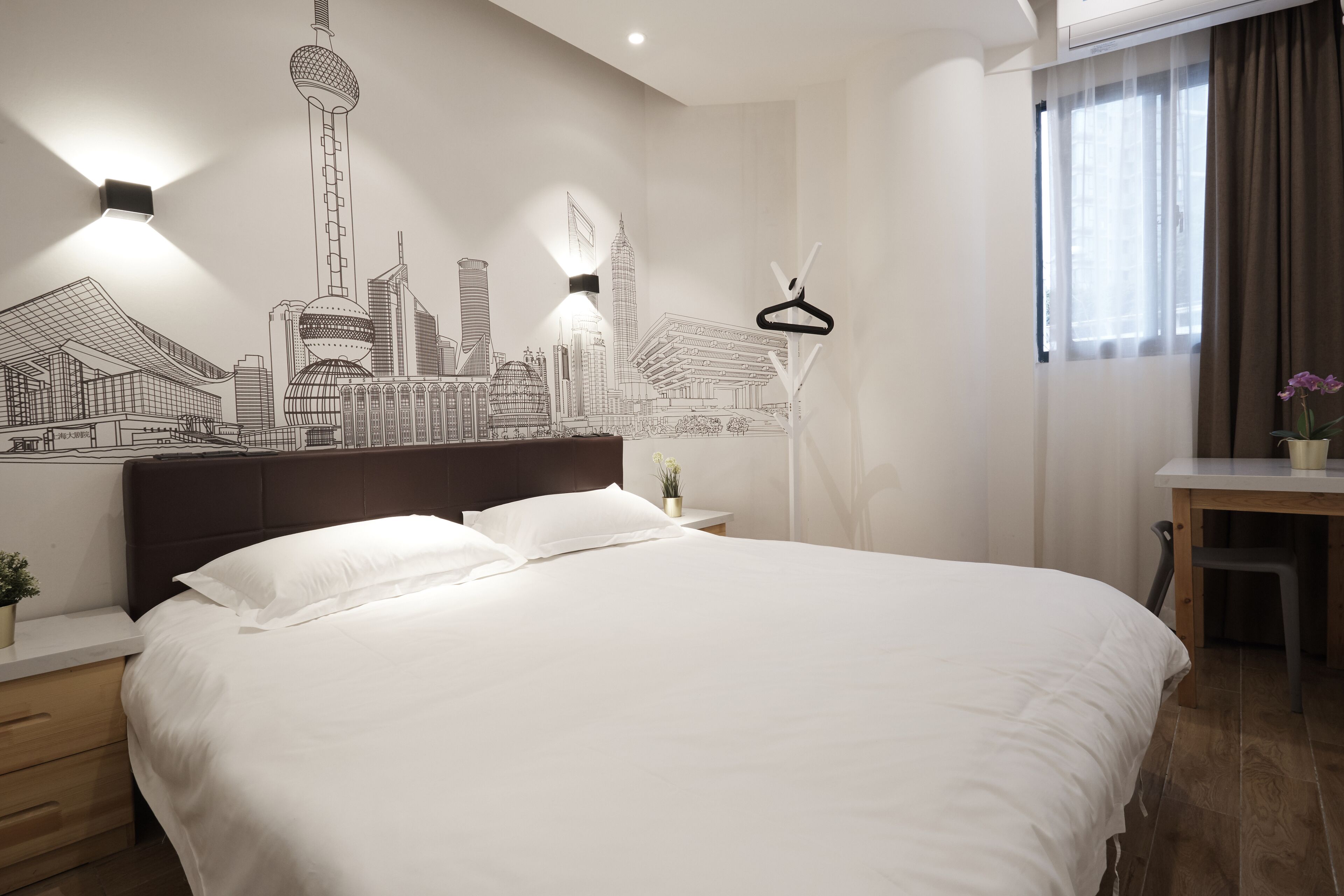 A modern hotel room with a large bed, a cityscape mural on the wall, a bedside table with a lamp, and a window with curtains.