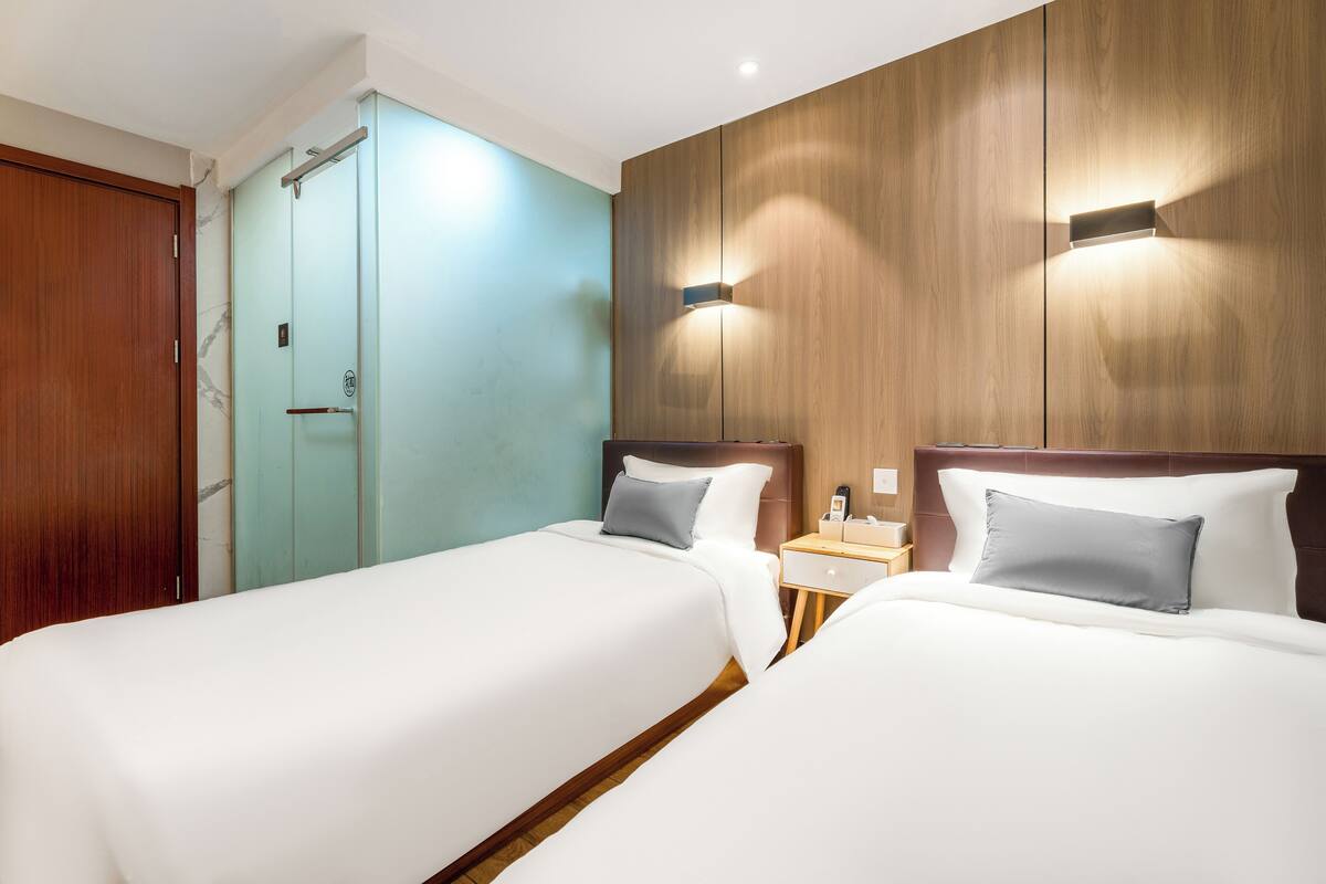 A hotel room with two beds, a wooden wardrobe, a glass shower enclosure, and a nightstand with a lamp.