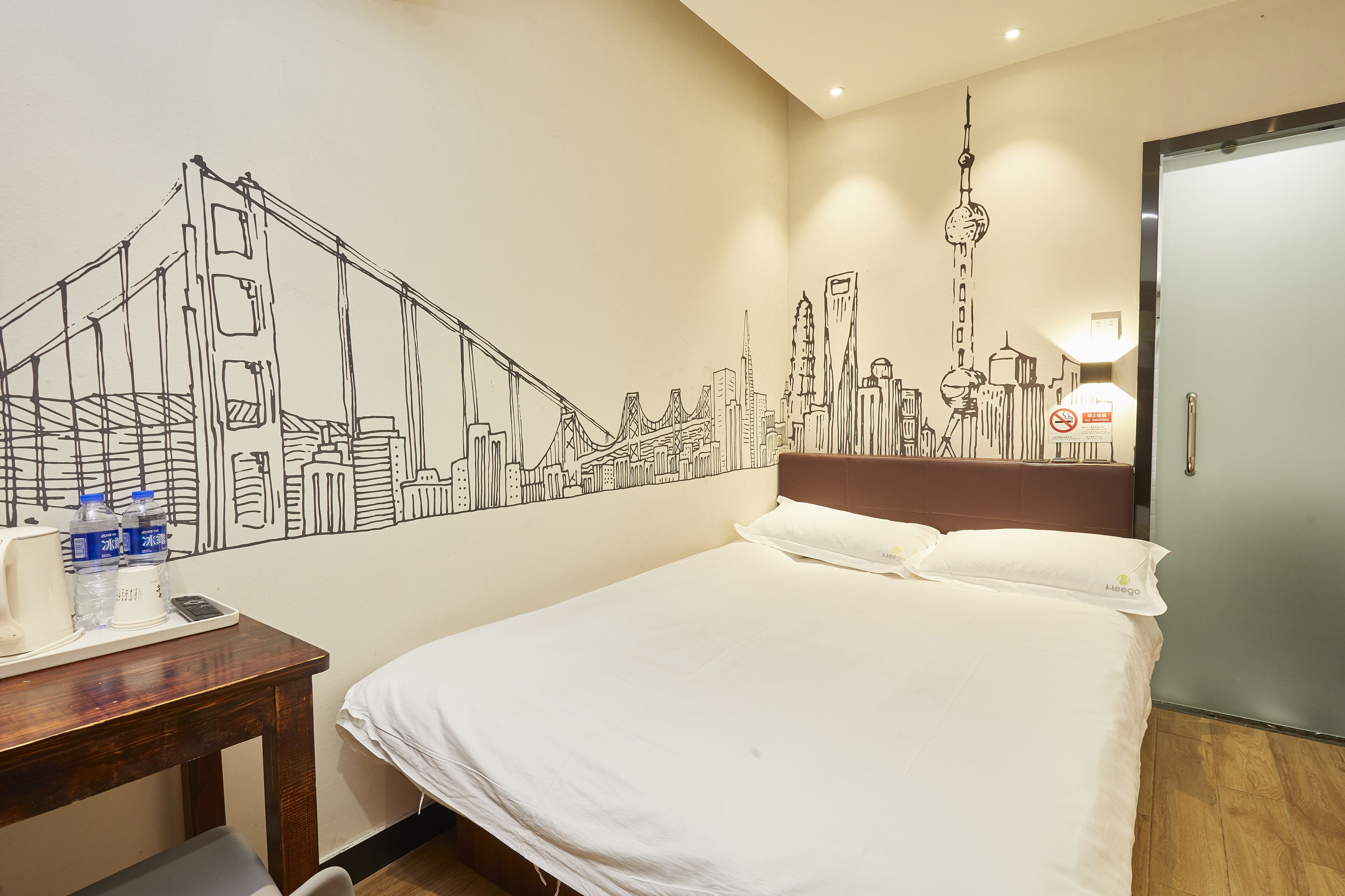 A hotel room with a bed, a desk with a phone and water bottles, and a wall mural of a city skyline.