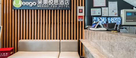 Zona de recepción con un cartel que indica Meego Yes Hotel, un panel de listones de madera, un mostrador de recepción con computadoras y un extintor.