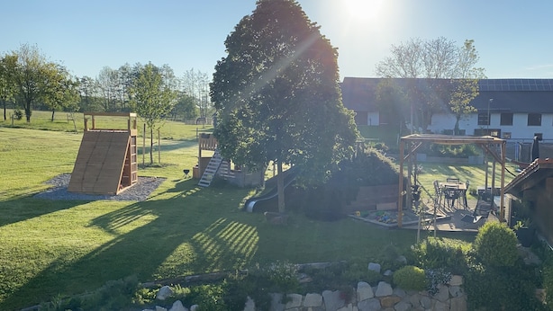 A grassy outdoor area with a wooden play structure, a stone wall, and a large tree. - Kramerhof (Taufkirchen)