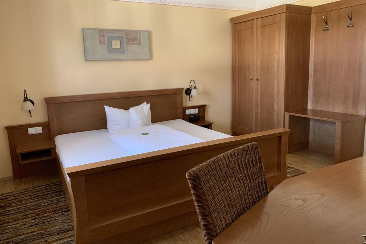Habitación de hotel con cama de madera, mesita de noche con lámpara, armario y una mesa de comedor.