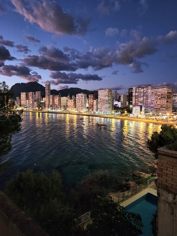 A nighttime cityscape with illuminated buildings reflecting on a river, a mountain in the background, and a pool area in the foreground.