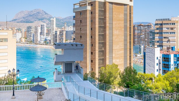 A coastal cityscape with modern buildings, a clear blue sea, and a mountainous backdrop.