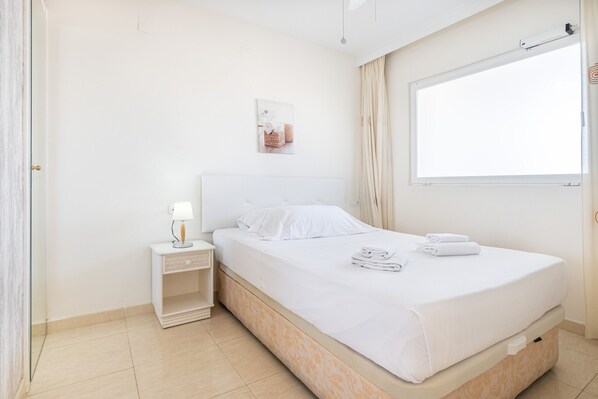 Una habitación de hotel ordenada con una cama individual, una mesita de noche con lámpara, un estante pequeño y una ventana con cortinas.
