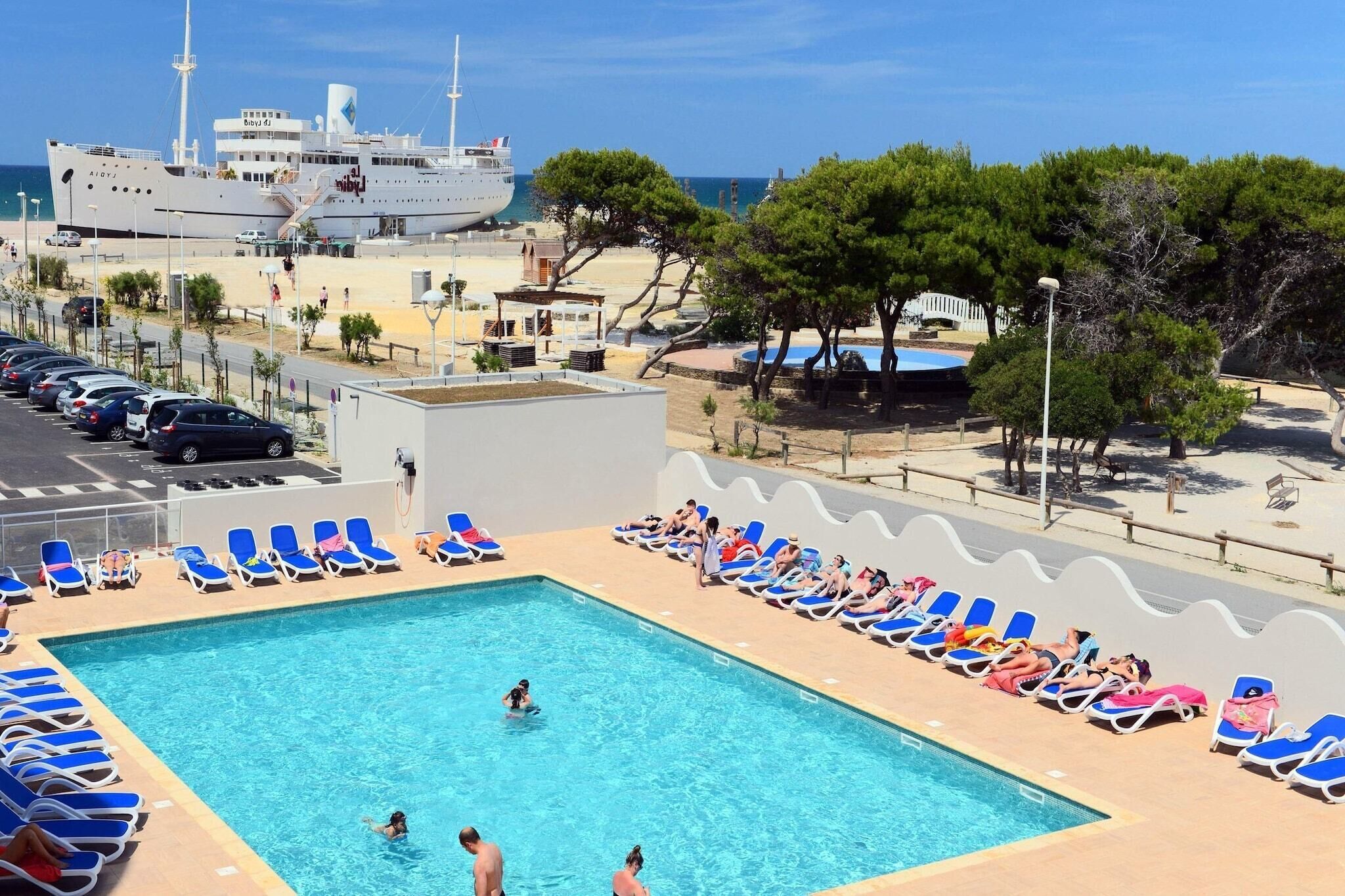 Une piscine avec des chaises longues, un espace de stationnement et un bâtiment en arrière-plan.