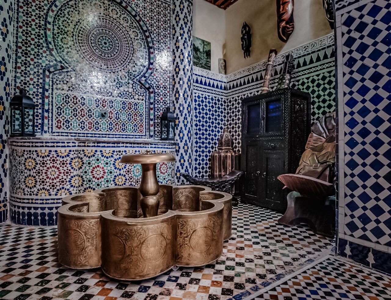 A traditional Moroccan interior with intricate tile work, a central water fountain, and a checkered floor pattern.