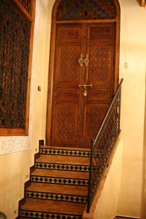 A wooden door with intricate carvings and a brass lock, flanked by a staircase with patterned tiles and a metal railing.
