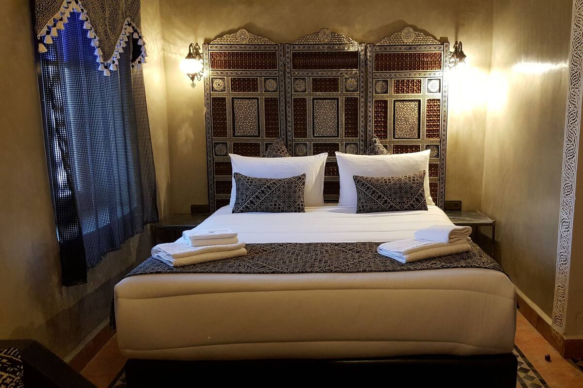 A large bed with a headboard featuring intricate patterns, two bedside tables, and a wall-mounted light fixture.