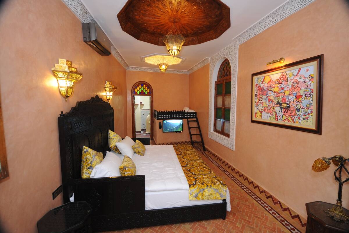 A bedroom with a large bed, wall-mounted lights, a wooden headboard, a framed picture, and a decorative ceiling.