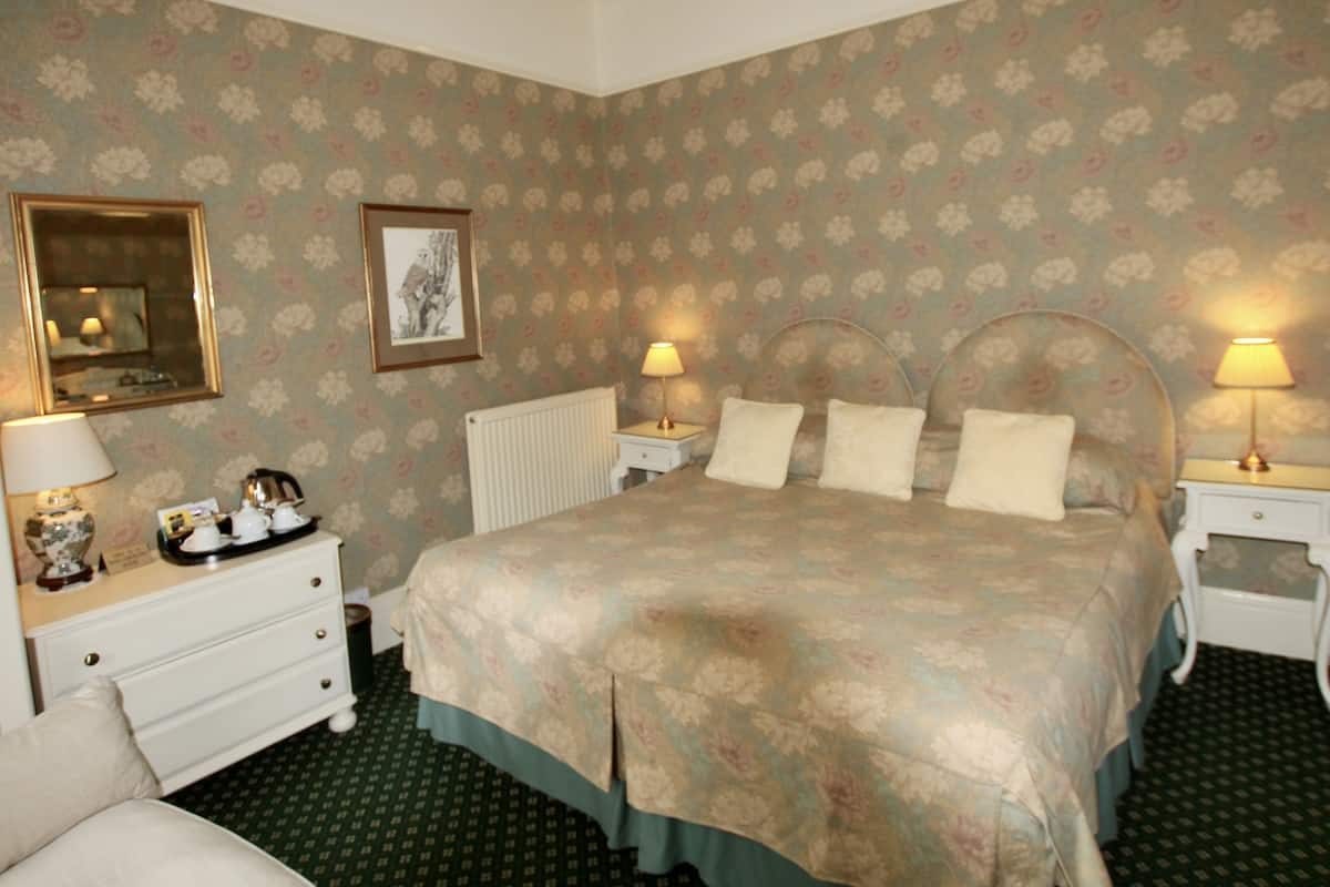 A bedroom with a floral-patterned wallpaper, a bed with a headboard, two bedside tables with lamps, a dresser, and a radiator.