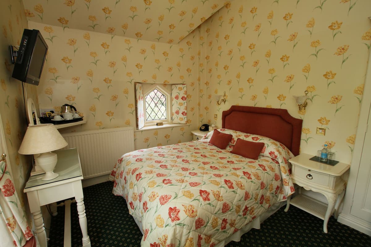 A bedroom with a floral-patterned bedspread, a television mounted on the wall, a bedside table with a lamp, and a window with a decorative grille.
