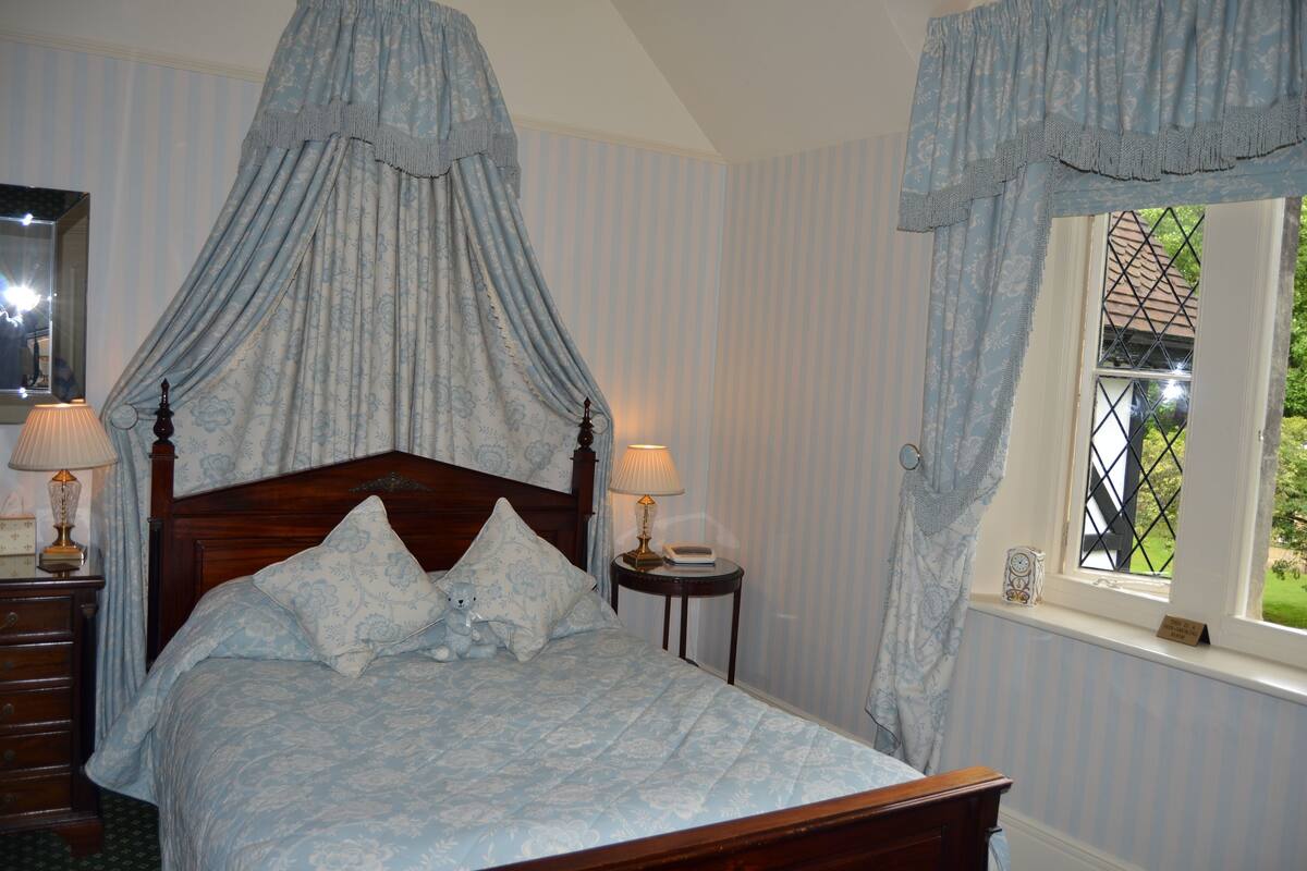 A four-poster canopy bed with floral bedding, two bedside lamps, and a window with a view of greenery.