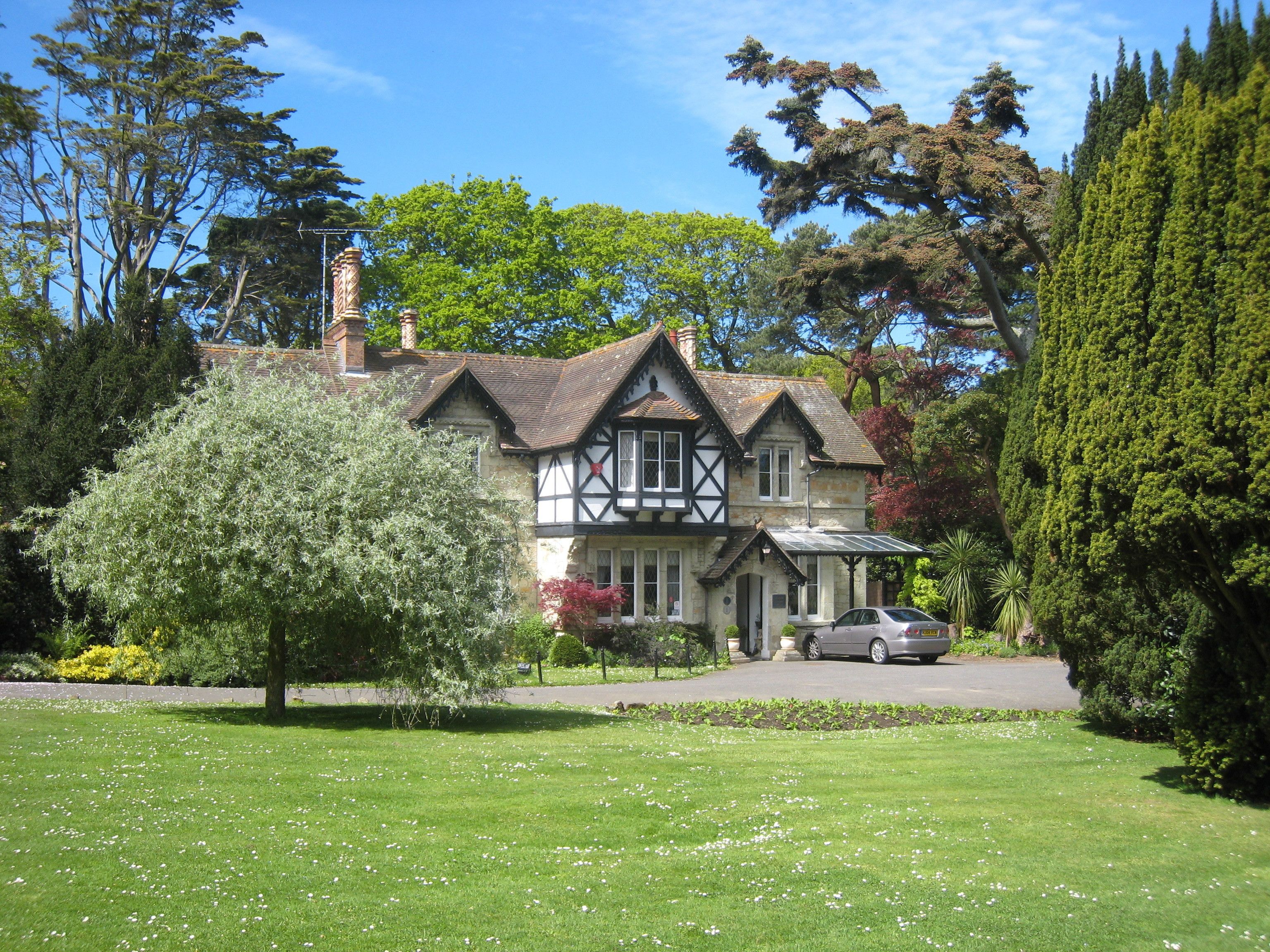 A traditional Tudor-style house with a stone facade, a steeply pitched roof, and a well-maintained lawn with white flowers.