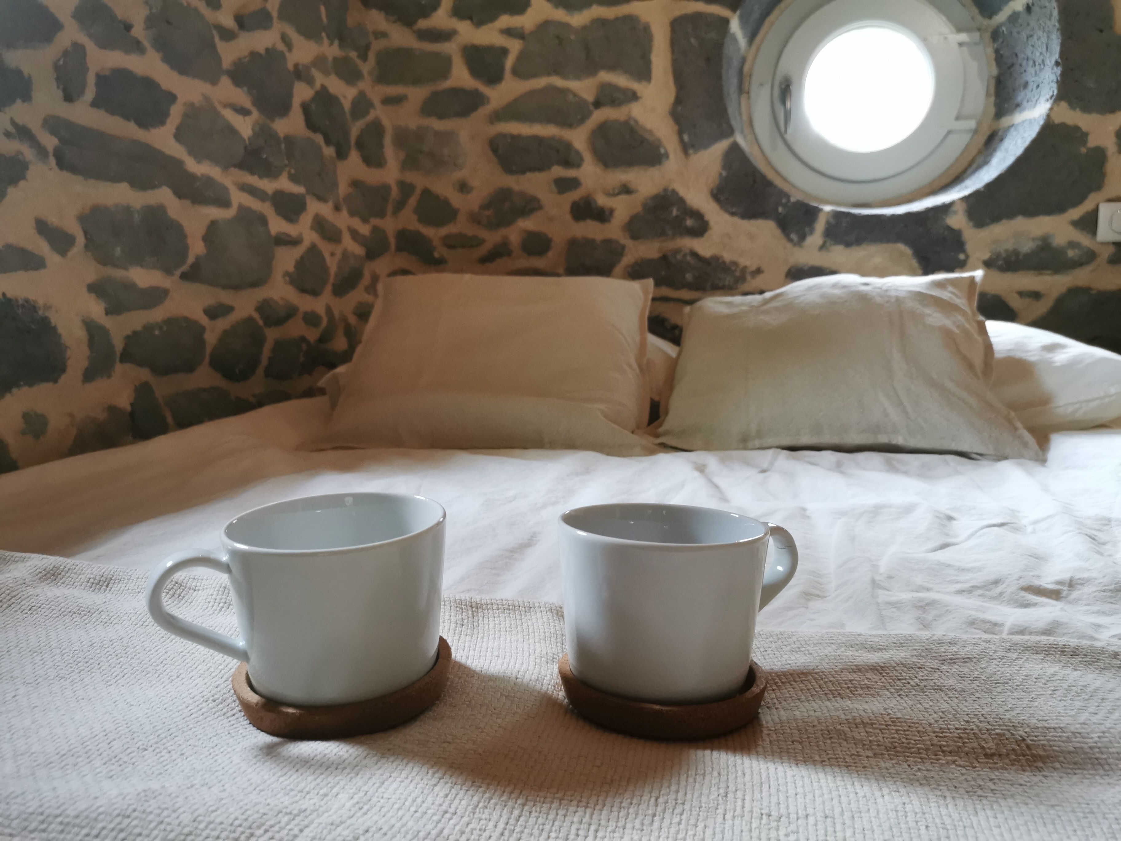 A bed with two white mugs on coasters, a stone wall, and a round window.