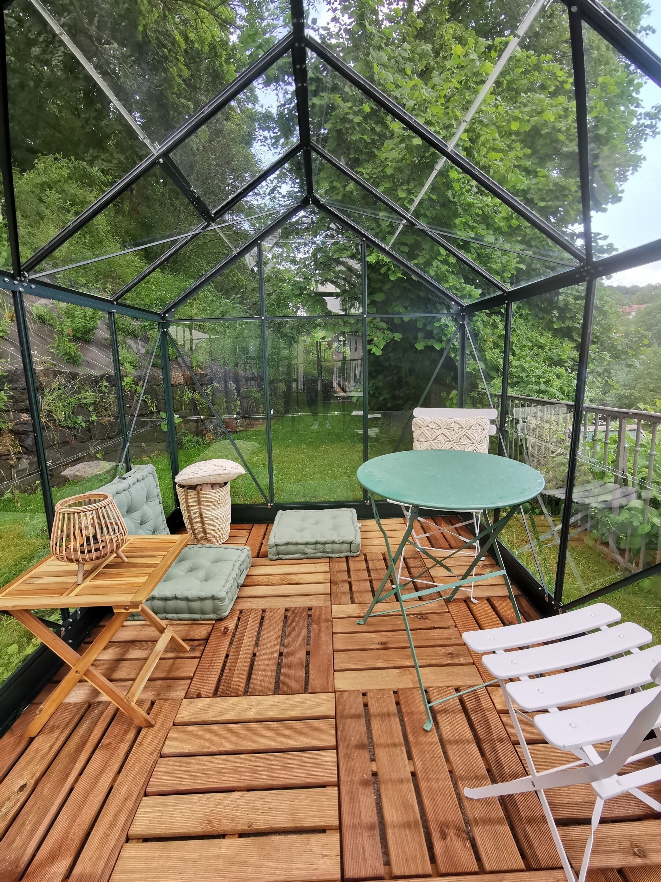 A greenhouse with a wooden deck, a table, chairs, cushions, and a wicker basket.