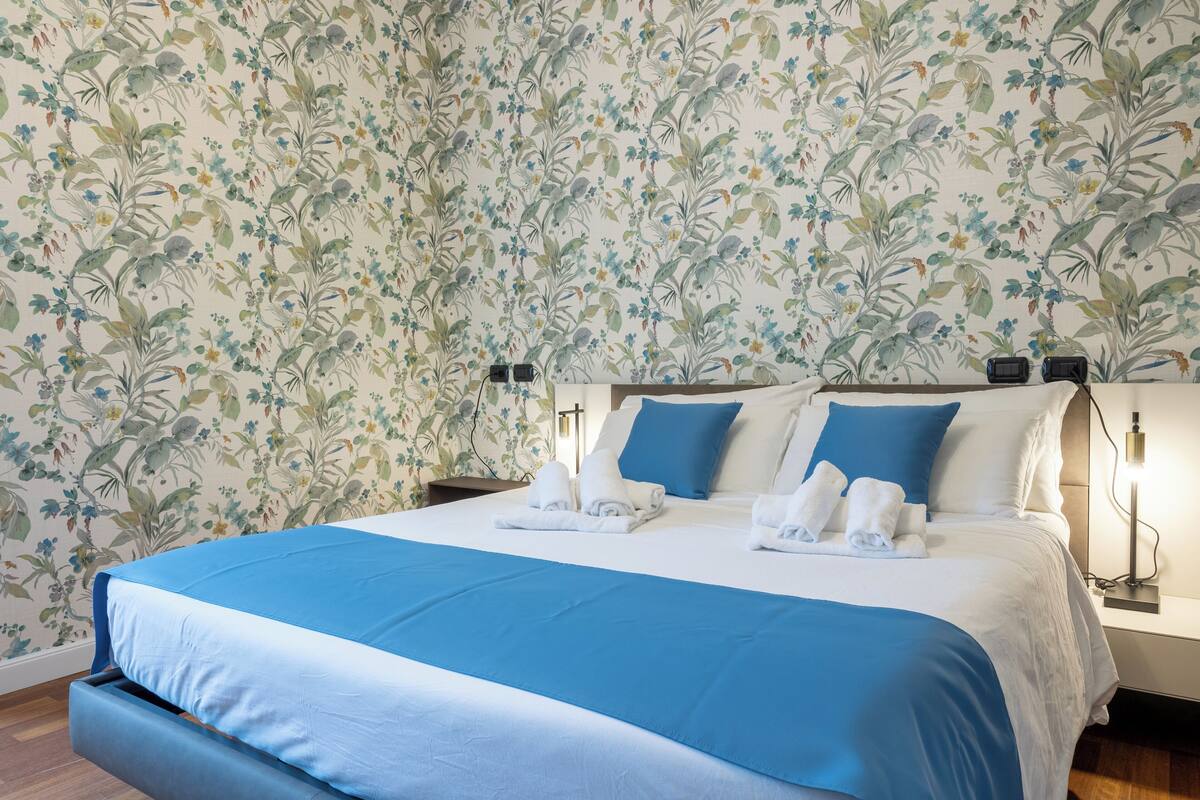 A neatly made bed with blue and white bedding, two bedside lamps, and a floral wallpaper pattern.