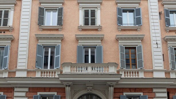 Un edificio de varios pisos con un estilo arquitectónico uniforme, con ventanas regularmente distribuidas con contraventanas, un balcón central con barandilla y molduras decorativas.