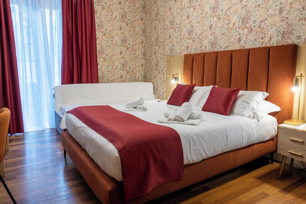 A neatly made bed with white linens and red accents, a wooden nightstand with a lamp, and a floral wallpaper pattern.