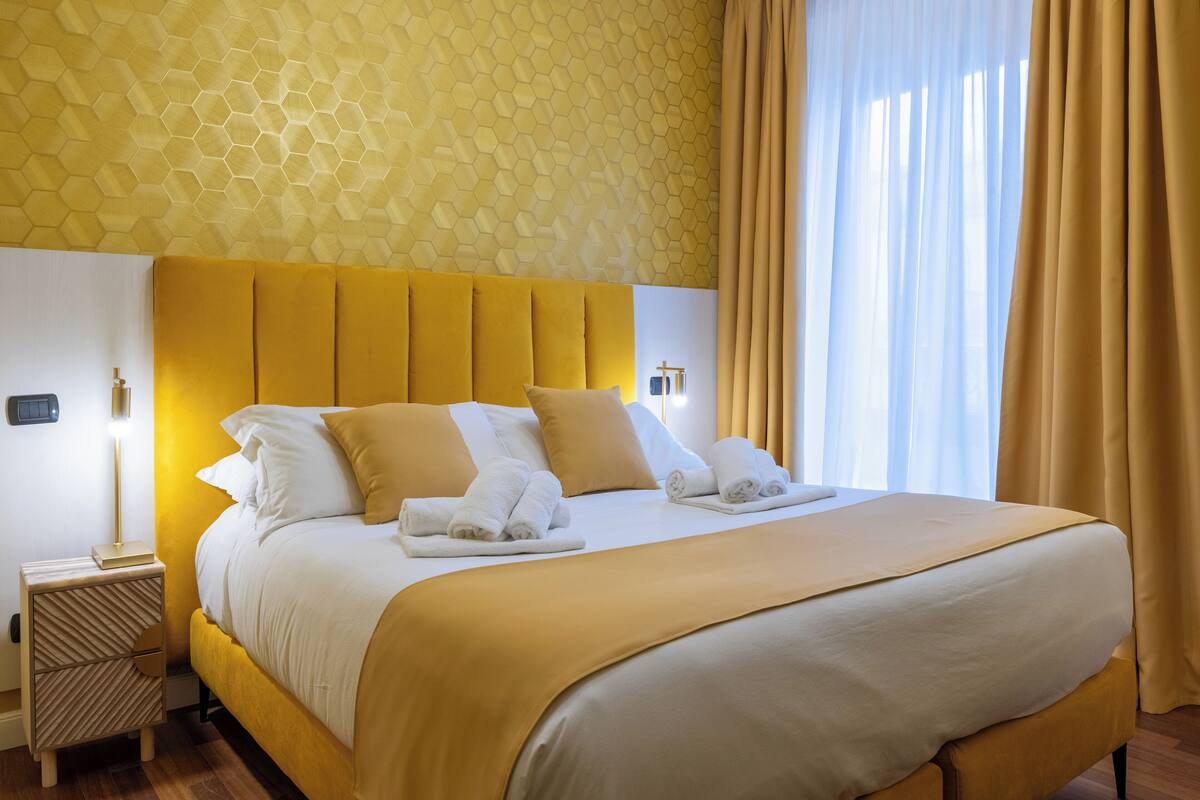 A hotel room with a yellow hexagonal wall pattern, a large bed with white linens and a yellow headboard, a bedside table with a lamp, and a window with curtains.