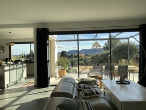 A modern living room with a sofa, a dining area, and a view of mountains through large windows.