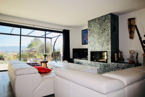 A modern living room with a white leather sofa, a fireplace, and a large window offering a view of the outdoors. - Superb contemporary villa with private pool (Saint-Rémy-de-Provence)