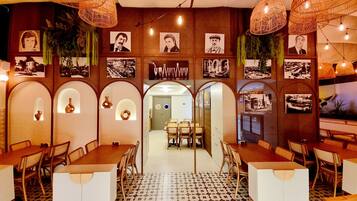 A restaurant with a central dining area, multiple tables, and chairs. There are wall-mounted picture frames and hanging lights.