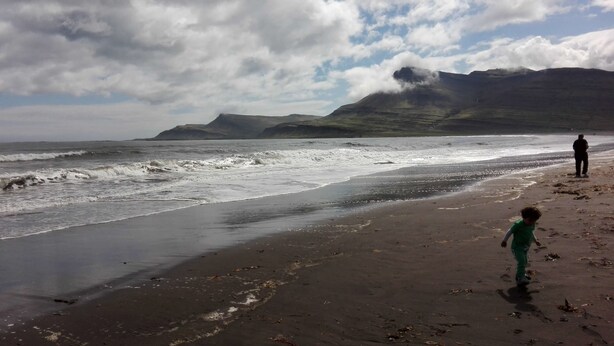 A beach with waves, a child, and a person standing on the sand. - Hótel Stadarborg (Fjardabyggd)