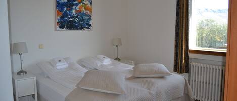 Una camera da letto con un letto, due comodini, una finestra con tende, un termosifone e un quadro appeso al muro.