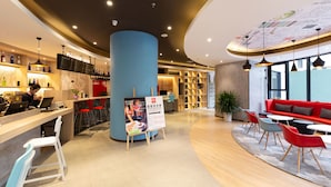 Een modern café met een bar, zitplaatsen met rode en witte stoelen, en een grote blauwe pilaar.