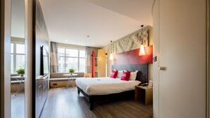 Een moderne hotelkamer met een groot bed, een nachtkastje, een raam met gordijnen, een klein tafeltje met een plant en uitzicht op de stad.