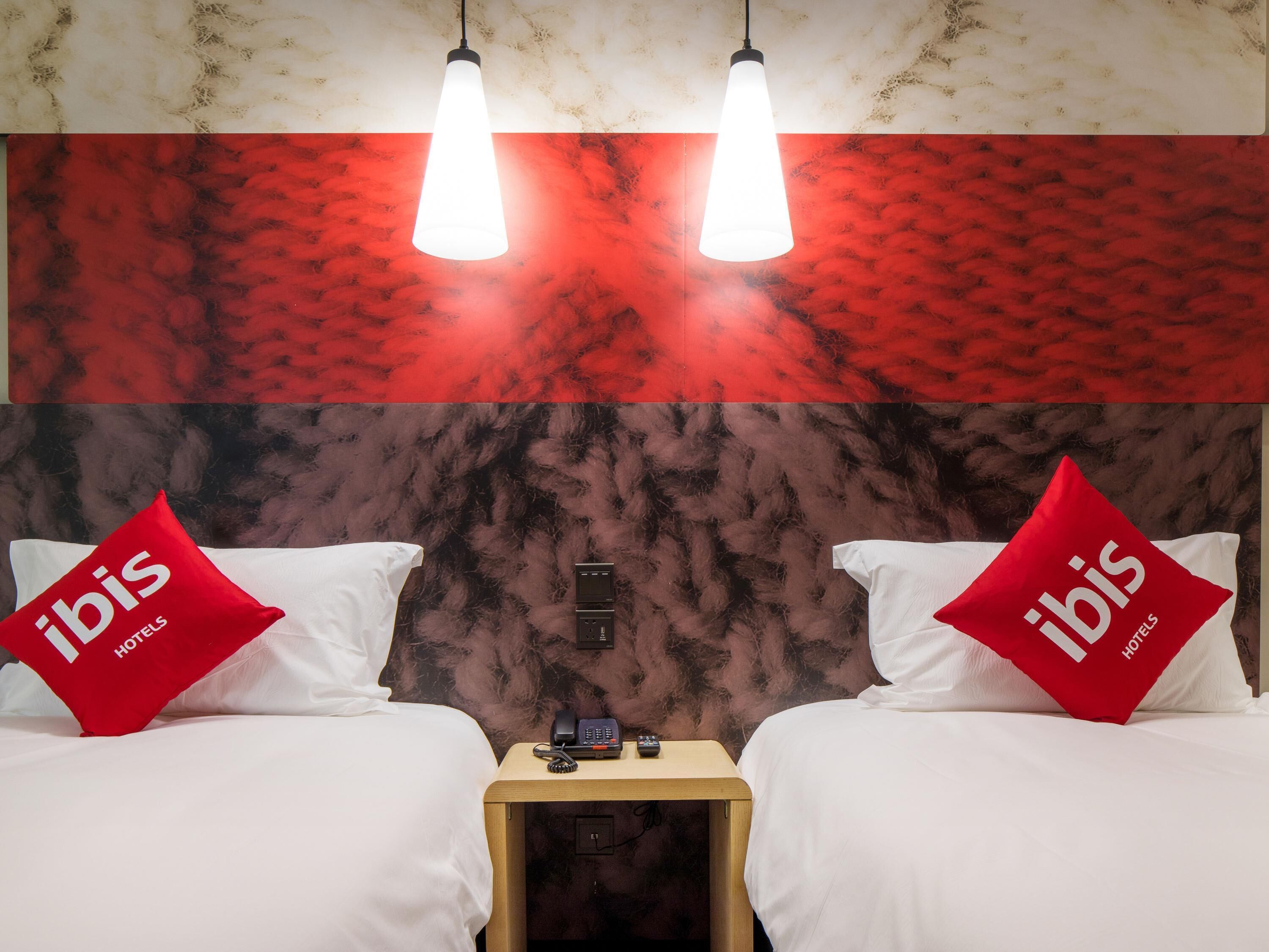 A hotel room with two beds, each with a red pillow labeled 'ibis Hotels'. There are two pendant lights hanging above the beds, and a wooden nightstand with a telephone and a remote control between the beds.