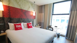 A hotel room with a large bed, two red pillows with "ibis Hotels" branding, a nightstand with a phone, a small table with a lamp, and a view of the city through a large window. - Ibis Hangzhou Future Sci-tech City Hotel (Hangzhou)