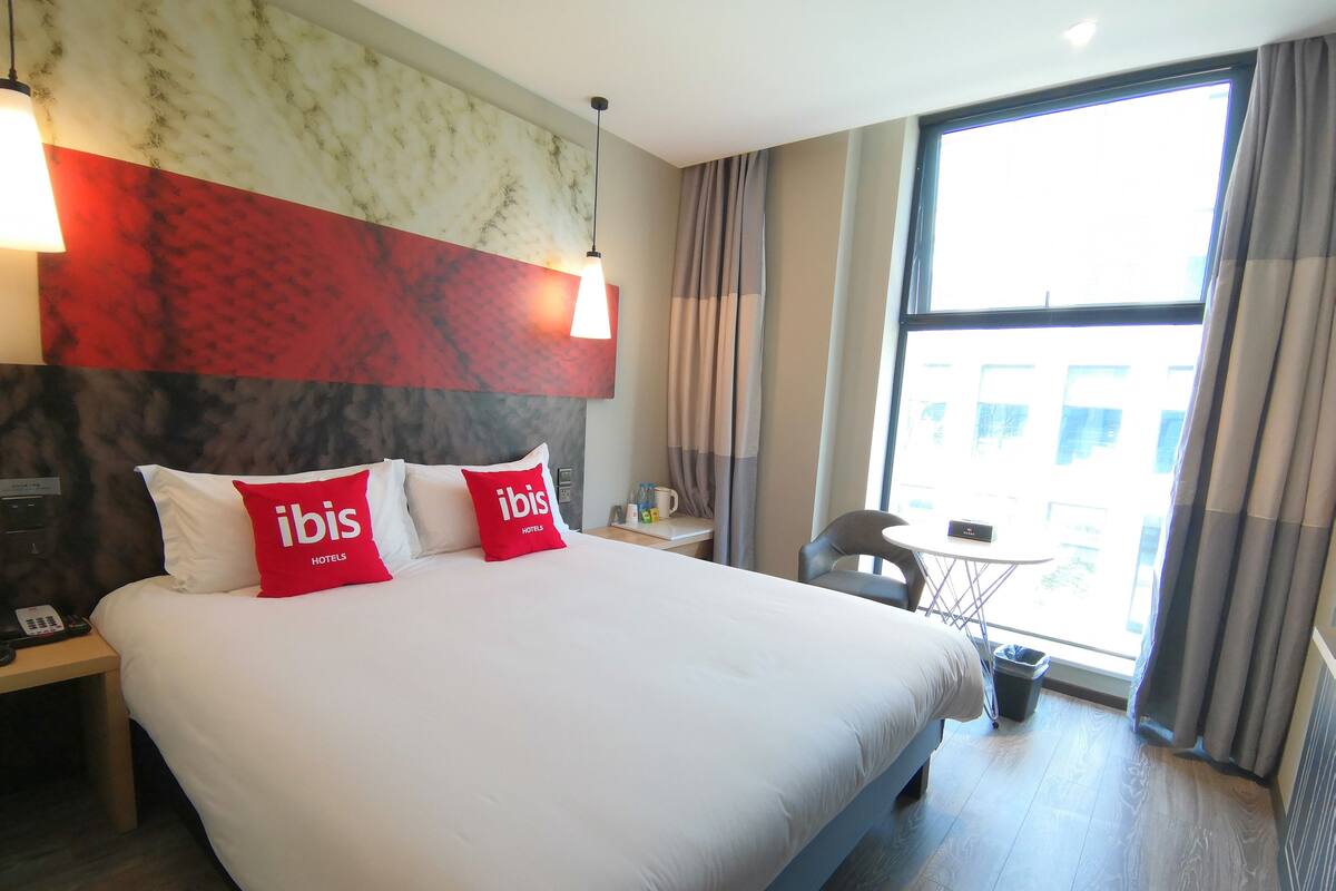 Quarto de hotel com uma cama grande, duas almofadas vermelhas com a marca 'ibis Hotels', uma mesa de cabeceira com um telefone, uma pequena mesa com um abajur e vista para a cidade através de uma grande janela.