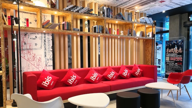 A modern hotel lobby with a red sofa, white chairs, and black stools. There are bookshelves with books and a whiteboard with handwritten notes. - Ibis Hangzhou Future Sci-tech City Hotel (Hangzhou)