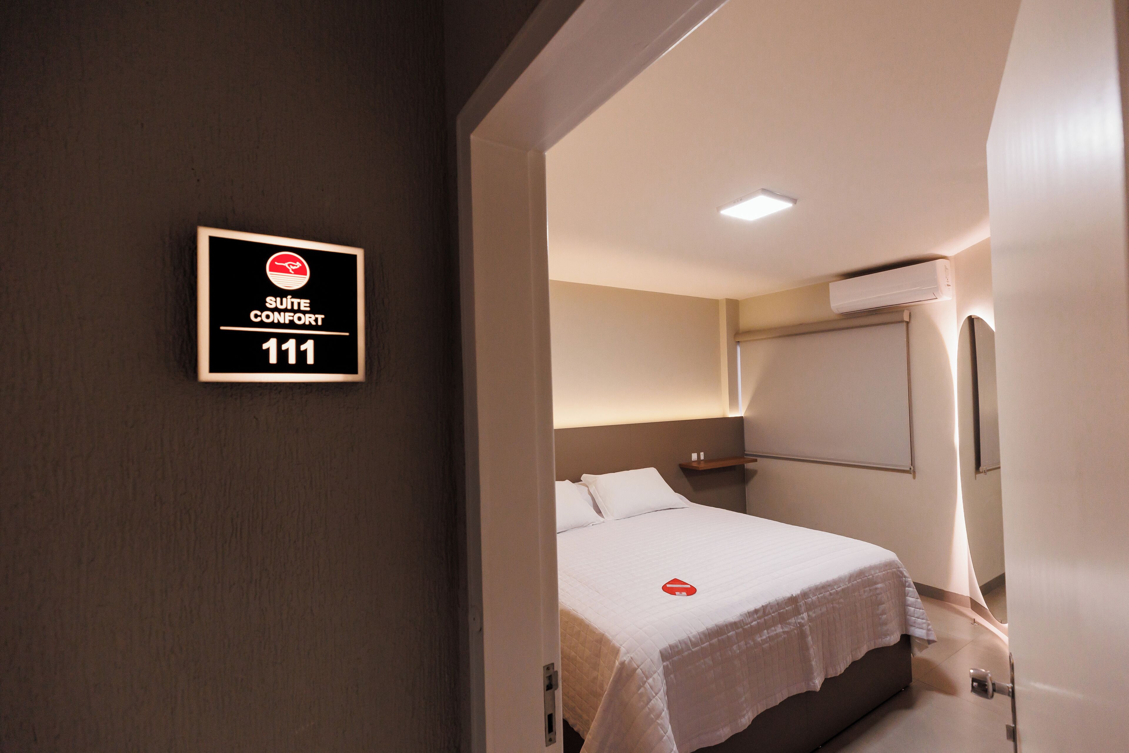 A hotel room with a bed, a nightstand, and a wall-mounted air conditioning unit. A sign indicates the room number as 111.