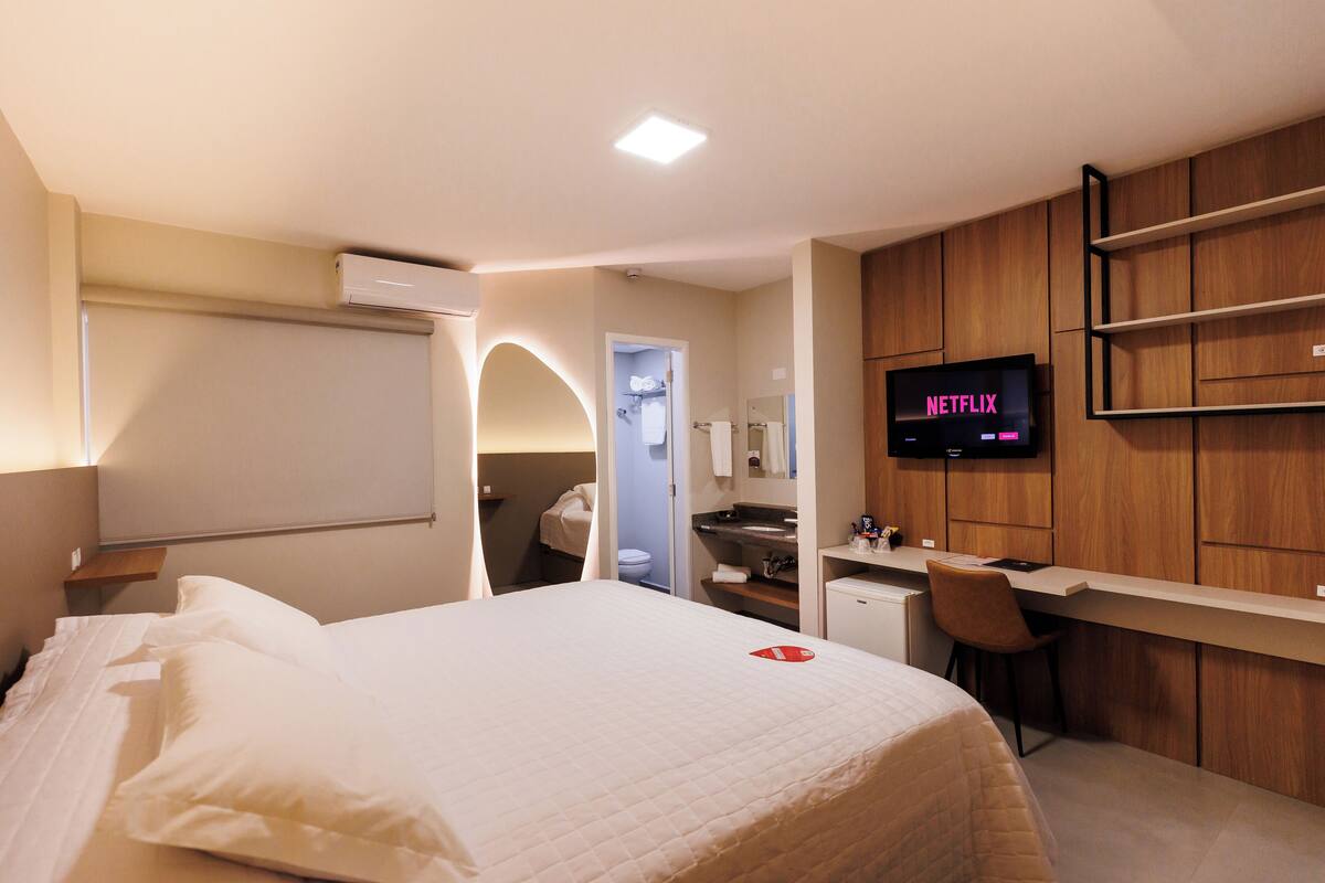 Une chambre d’hôtel avec un lit, un bureau, un téléviseur affichant le logo « NETFLIX », et une salle de bain visible à travers une arche.