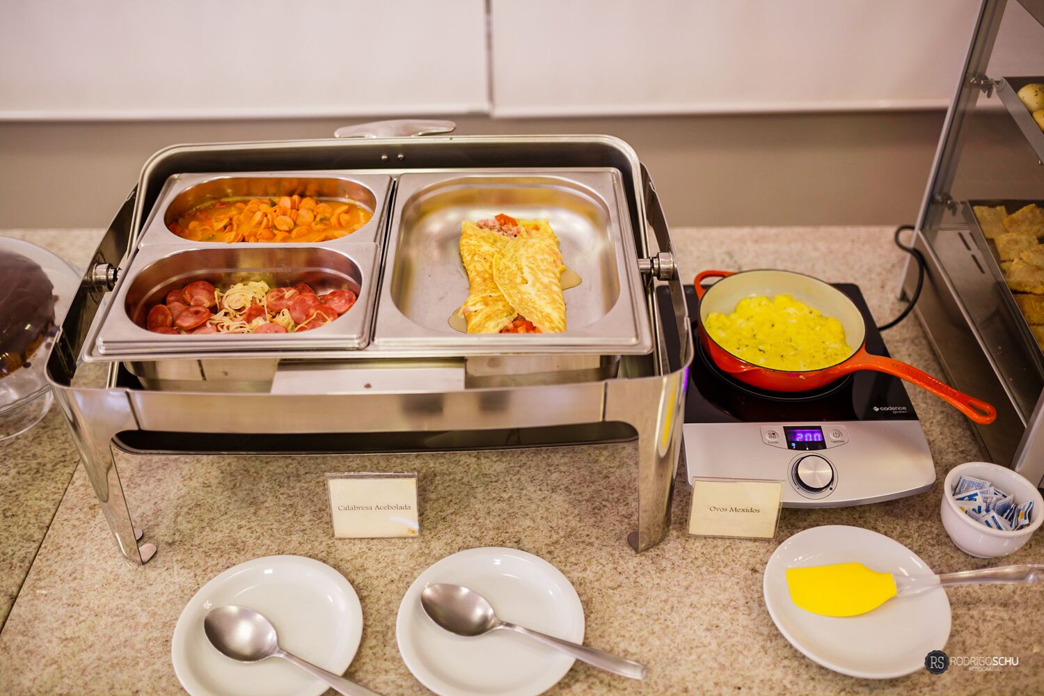 Montaje de bufé con platos identificados, incluyendo Calabresa Acebollada, Huevos Revueltos y una olla de huevos revueltos.
