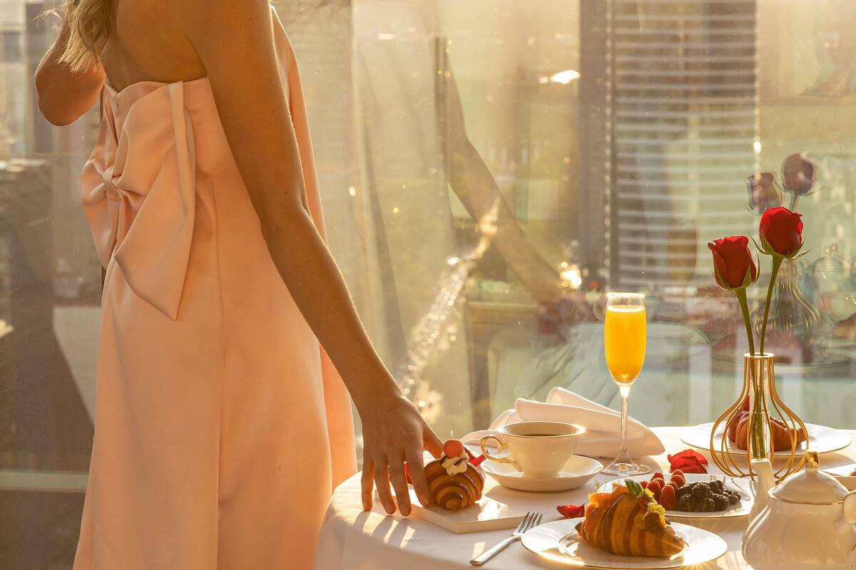 Eine Frau in einem hellrosa Kleid steht an einem Tisch mit Gebäck, einem Glas Orangensaft und einer Vase mit roten Rosen.