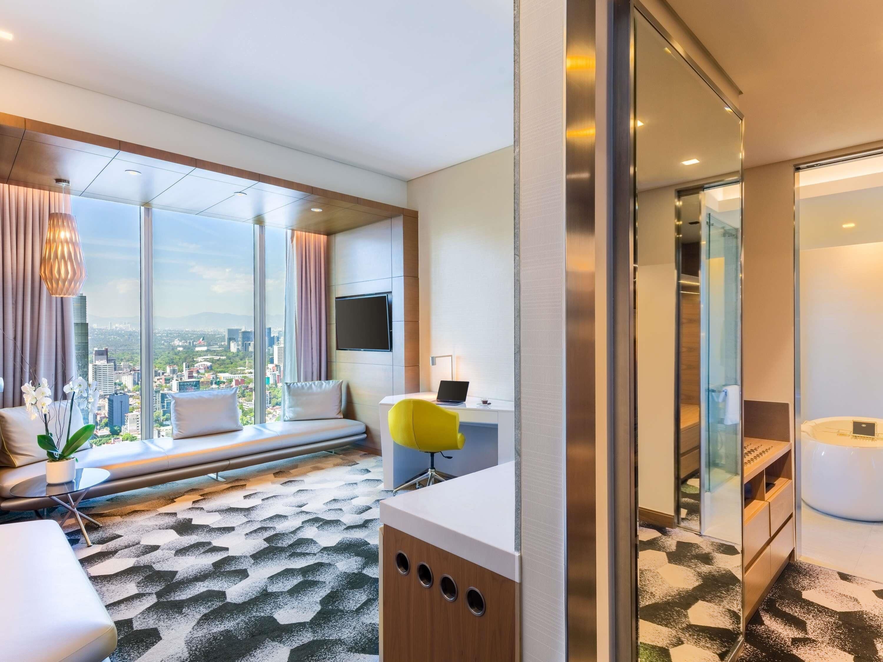 A modern hotel room with a large window, a bed, a desk, and a bathroom.