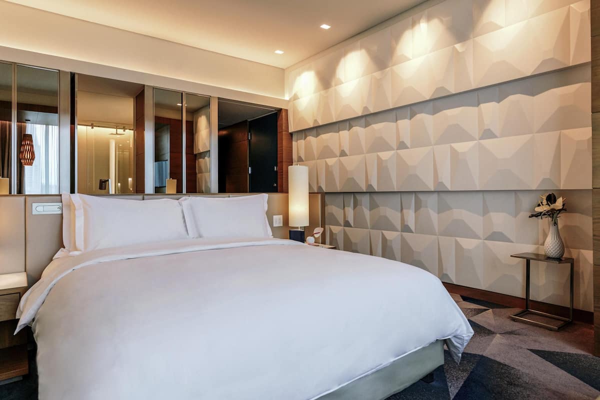 Une chambre d’hôtel moderne avec un grand lit, un miroir, une table de chevet et une tête de lit ornée d’un motif géométrique.